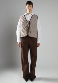 Quiltet vest i beige med diamantmønster og brune accenter, båret over en hvid skjorte, parret med brune bukser og sorte spidse sko.