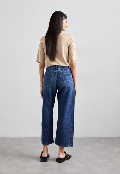 Beige t-shirt, losse fit, gecombineerd met hoge taille, wijdvallende donkerblauwe jeans met twee achterzakken met een zigzagpatroon. Zwarte open schoenen.