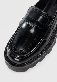 Must musta nahk loafers brogue perforatsioonidega, must paks tald ja hõbedane DKNY logo nahkriidel ülevalt.