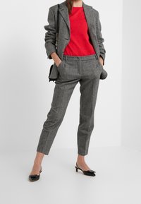 Kvinde iført en grå stribet buksedragt med en rød struktureret top, sorte slingback hæle og en sort skuldertaske, der står med hænderne i lommerne.