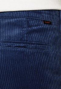 Tmavě modré manšestrové kalhoty s vertikálními rýhami, vybavené boční kapsou a malou černou etiketu s červeným nápisem.