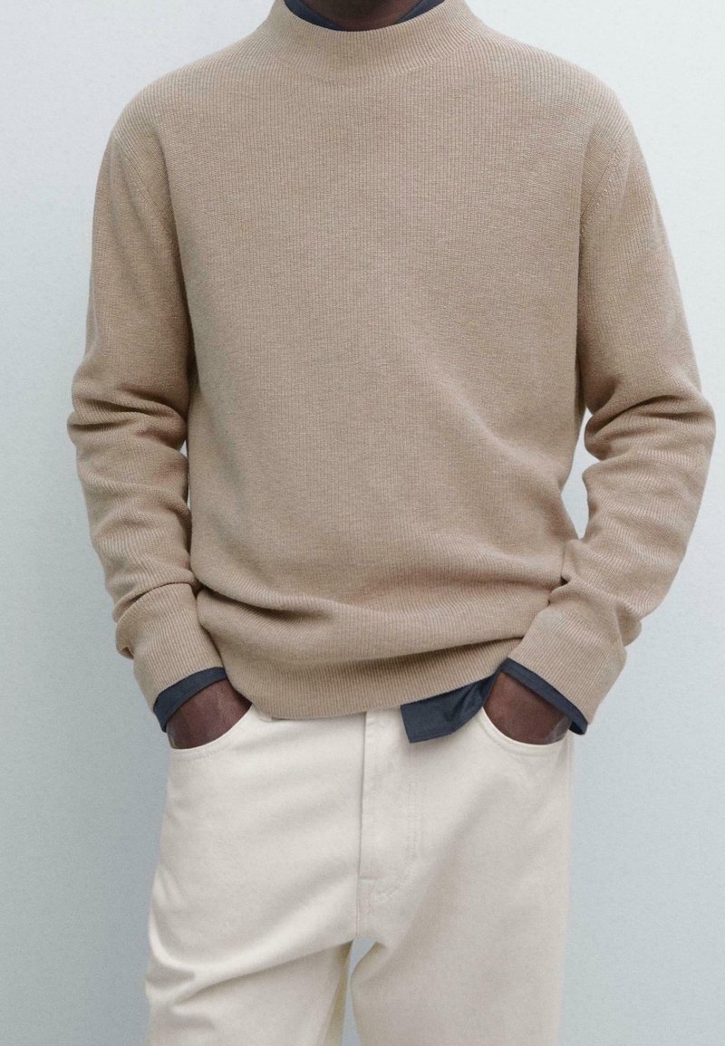 Homme portant un pull beige à côtes par-dessus une chemise sombre, les mains dans les poches d’un pantalon couleur crème, debout sur un fond uni.