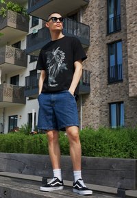 Svart grafisk T-skjorte med engel-design, denimshorts, hvite sokker, og svarte Vans-sneakers. Utendørs setting med moderne leilighetsbakgrunn.