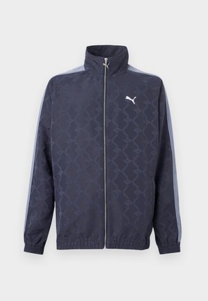 Jachetă cu fermoar închisă pe culoarea albastru închis, cu un model geometric subtil, dungi bleu pe mâneci, manșete și tiv elastice, și logo alb Puma pe piept.