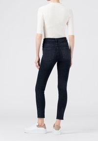 Jeans ajustados de cintura alta de color azul oscuro con bolsillos traseros y una textura suave, combinados con una blusa de manga corta de color claro y zapatillas blancas informales.