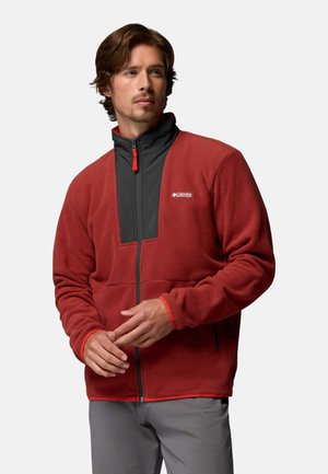 Mann trägt eine rote Fleecejacke mit schwarzem Brustpaneel und Reißverschluss, steht mit verschränkten Händen und schaut zur Seite.