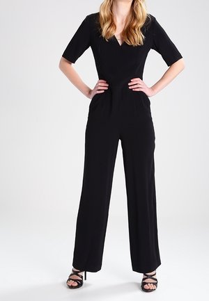 Schwarzer Overall mit kurzen Ärmeln, V-Ausschnitt, tailliertem Oberteil und weitem Hosenbein. Material wirkt glatt und leicht. Schwarze Riemchenschuhe.