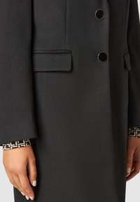 Detailní záběr rukou podél těla v tmavém texturovaném kabátu s klopovými kapsami a černými kulatými knoflíky přes vzorovanou černo-bílou košili.