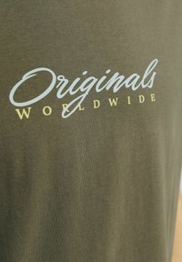Olivezelené bavlnené tričko s bielym a žltým textom na skripte "Originals Worldwide." Hladká textura, krátke rukávy, ležérny dizajn.