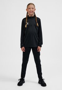 Junge Person steht, trägt schwarze Sportkleidung und hat geflochtene Haare, lächelt vor einem schlichten hellen Hintergrund.