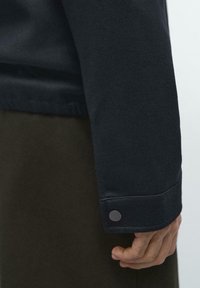 Rękaw ciemnoniebieskiego sweatshirtu z gładką teksturą, regulowany mankiet z zaokrąglonym metalowym zapięciem oraz prosty, elegancki design.