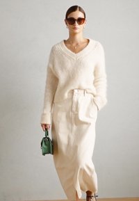 Beige knitted sweater with a V-neck, paired with a cream maxi skirt. The outfit includes a small green handbag as an accent detail.