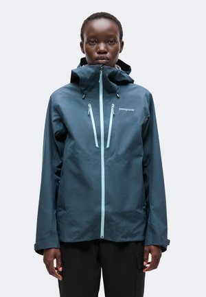 Personne portant une veste imperméable bleu foncé avec une capuche et des fermetures éclair bleu clair, debout devant un fond clair uni.