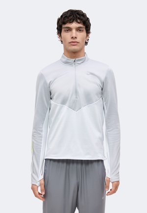 Uomo che indossa un top a maniche lunghe grigio chiaro con mezza cerniera e fori per i pollici e pantaloni sportivi grigio scuro, in piedi rivolto verso il fronte.