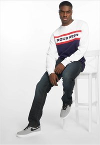 Hvit hettegenser med røde og marineblå detaljer, prydet med "ROCA WEAR" grafikk. Mørkeblå jeans og grå Nike-sko. Mann sitter på en hvit krakk.