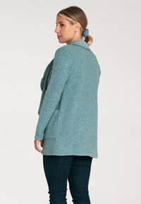 Jasnoniebieski kardigan o teksturowanym wykończeniu, z kołnierzem typu szal i kieszeniami z przodu, noszony na ciemnych dżinsach. Tkanina wydaje się miękka i przytulna.