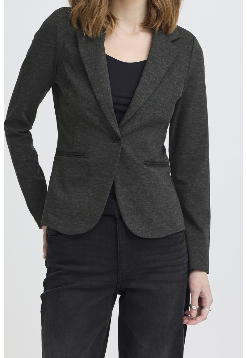Femme portant un blazer gris foncé ajusté avec un bouton unique sur un haut noir et un pantalon noir, debout avec une main sur la hanche.