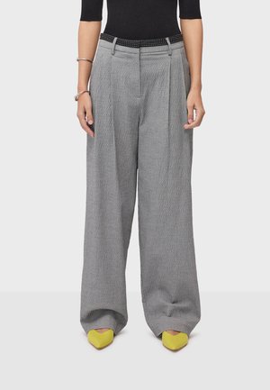 Žena u širokim hlačama visokog struka sive teksture s crnim remenom, crnom majicom, srebrnim narukvicama i žutim špicastim cipelama.