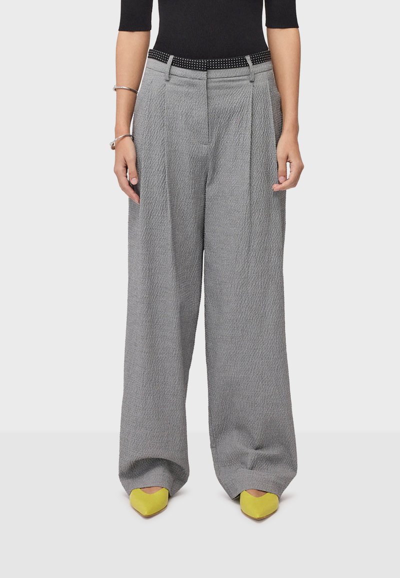 Femme portant un pantalon gris texturé taille haute à jambes larges avec une ceinture noire, un haut noir, des bracelets argentés et des chaussures pointues jaunes.