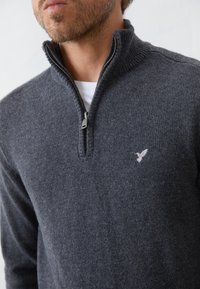 Dunkelgrauer Strickpullover mit Reißverschluss; aus gestricktem Stoff gefertigt; verfügt über einen hohen Kragen und ein kleines, auf der Brust gesticktes Logo. Glatte Textur, lockere Passform.