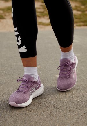 Gros plan sur des jambes portant des leggings noirs et des chaussettes blanches, marchant sur le trottoir avec des chaussures de course violet clair avec des lacets noués.