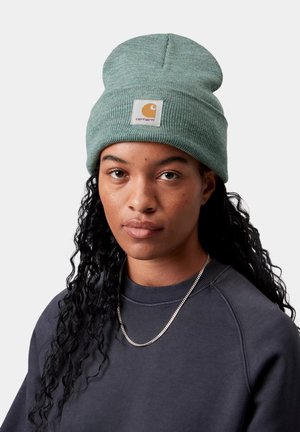 Persona con largo cabello rizado negro, usando un gorro de lana verde de Carhartt, sudadera gris oscuro y collar de cadena plateada, sobre un fondo claro.