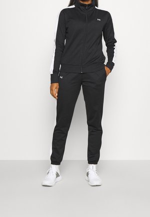 Persoon gekleed in een zwart-wit Under Armour trainingspak met een ritsvest en bijpassende broek, gecombineerd met witte sneakers, staand tegen een witte muur.