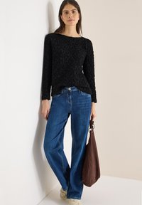 Černý svetr s texturou a dlouhými rukávy, kombinovaný s modrými džínami rovného střihu. V pravé ruce je držena hnědá semišová taška.