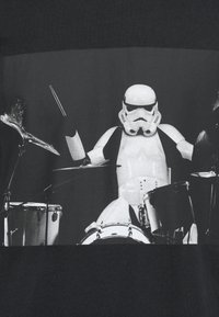 T-shirt preta com uma ilustração de um Stormtrooper a tocar bateria, com equipamento de bateria detalhado representado em tons de cinza.