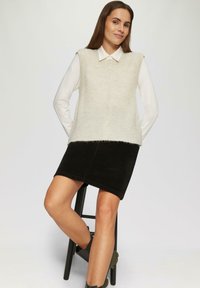 Lichtbeige gebreid vest met knopen over een witte longsleeve, gecombineerd met een zwarte corduroy rok en groene enkellaarzen.