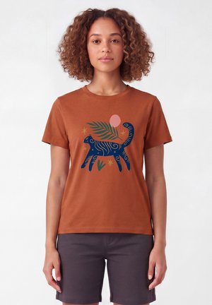 Vrouw met krullend haar, draagt een bruin T-shirt met een blauw kat- en plantmotief, gecombineerd met donkergrijze knielange shorts, staande tegen een witte achtergrond.