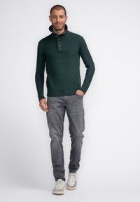 Donkergroen gebreide pullover met een geribde kraag, voorzien van twee knopen, gecombineerd met grijze jeans en lichtgekleurde sneakers.