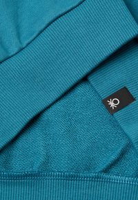 Sudadera verde azulado con puños acanalados y un cuerpo texturizado. Incluye una etiqueta negra con un logo en la parte inferior derecha. Detalle de tejido suave visible.