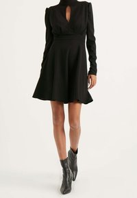 Robe noire à manches longues avec un col haut et un détail en forme de trou de serrure, corsage ajusté et jupe évasée. Portée avec des bottines noires.
