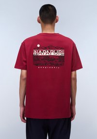 Camiseta de algodón roja con un diseño gráfico en la parte de atrás que presenta montañas, árboles y el texto "NAPAPIJRI geographic" encima de un círculo blanco.