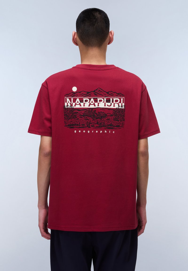 Camiseta de algodón roja con un diseño gráfico en la parte de atrás que presenta montañas, árboles y el texto "NAPAPIJRI geographic" encima de un círculo blanco.