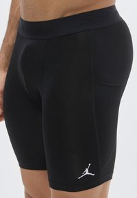 Μαύρο αθλητικό σορτς από λείο, ελαστικό υλικό. Διαθέτει φαρδιά, σκούρα ζώνη μέσης με το λογότυπο "JORDAN" και ένα λευκό γραφικό Jumpman.