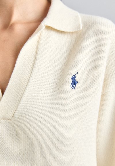 Krémový pletený polo svetr s V-výstřihem a krátkým límcem, s malým modrým vyšitým logem na levém hrudníku.