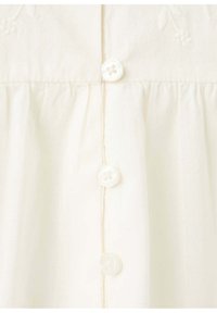 Gros plan d'un vêtement de couleur crème avec fermeture verticale à boutons comportant trois boutons blancs à quatre trous sur un tissu froncé.