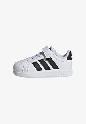 Sneaker bianca Adidas con strisce nere, lacci e una fascia in velcro, vista di lato su sfondo bianco.