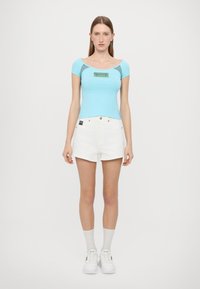 Light blue fitted top with a scoop neckline and short sleeves, featuring a graphic design. Paired with white denim shorts and white sneakers.
