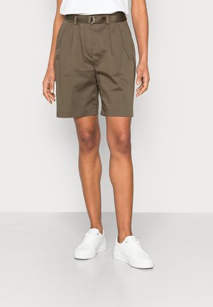 Personne portant un short ceinturé vert olive longueur genou, des baskets blanches et un haut blanc, debout sur un sol clair uni et un fond clair.
