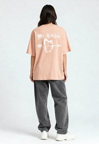 T-shirt surdimensionné rose clair avec un imprimé graphique blanc représentant un cœur avec une flèche, associé à un pantalon gris ample et des baskets blanches.