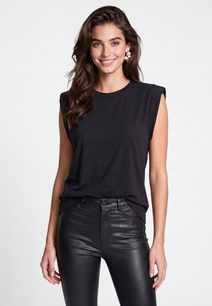 Young woman with long brown hair wears a sleeveless black top, black leather pants, and gold flower-shaped earrings, smiling, on white background.