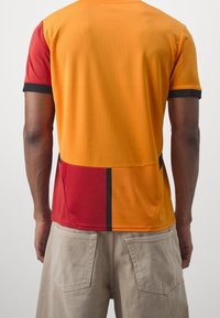 Camisa de manga corta con un cuerpo naranja, acentos rojos y ribete negro. Fabricada con una tela texturizada y un diseño de bloques de color.