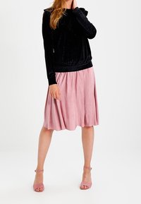 Svart velour långärmad topp med ribbad nederkant som är matchad med en rosa plisserad midikjol. Modellen bär matchande rosa klackar.