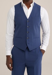 Námořnická modrá upravená vesta s hladkým povrchem, zapínání na pět knoflíků a jedna náprsní kapsa; v kombinaci s bílou košilí s dlouhým rukávem.
