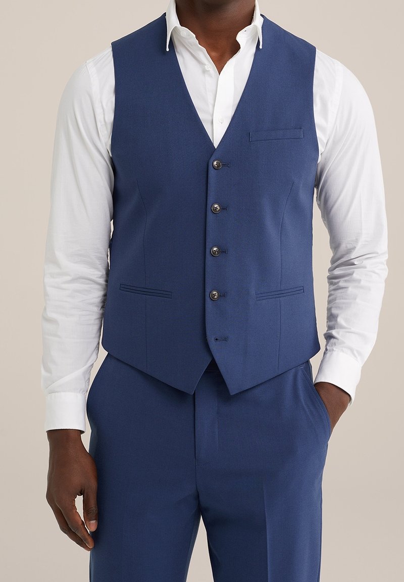 Námořnická modrá upravená vesta s hladkým povrchem, zapínání na pět knoflíků a jedna náprsní kapsa; v kombinaci s bílou košilí s dlouhým rukávem.