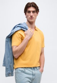 Jeune homme portant une chemise jaune à manches courtes et un jean bleu clair, tenant une veste en denim sur l'épaule, regardant devant lui.