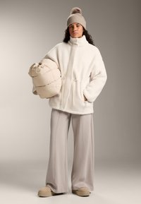 Krém színű polárdzseki magas gallérral, deréknál szűkített, oldalzsebekkel. Bézs színű puffadt táska húzózsinórral. Világosszürke, széles szárú nadrág és barna cipő.